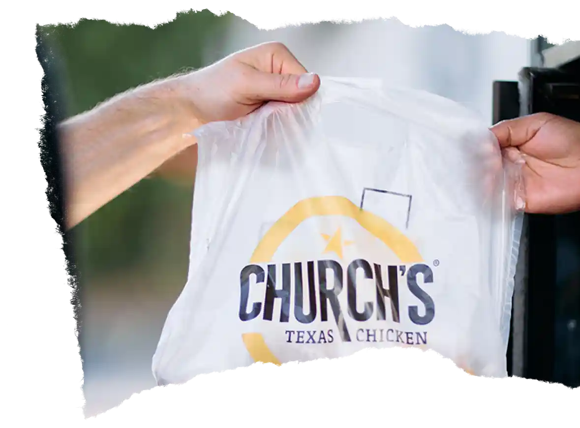 Hand passing a Church’s Texas Chicken take-out bag to another person.